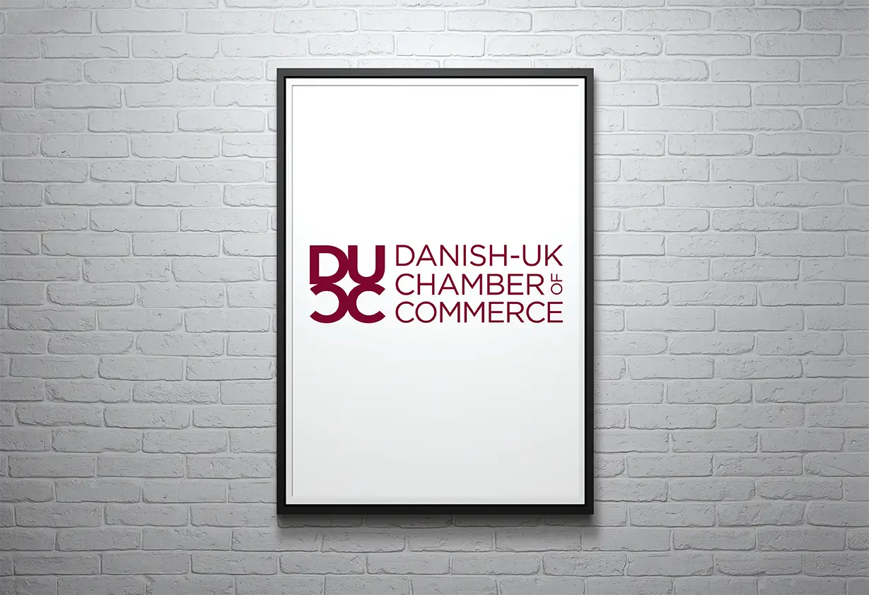 A framed poster displaying Danish-UK Chamber of Commerce logo on a white brick wall - originated by WarmRed